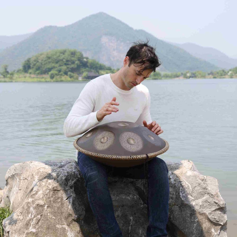 Handpan player using a Cosmoshandpan instrument beside a calm, scenic lake