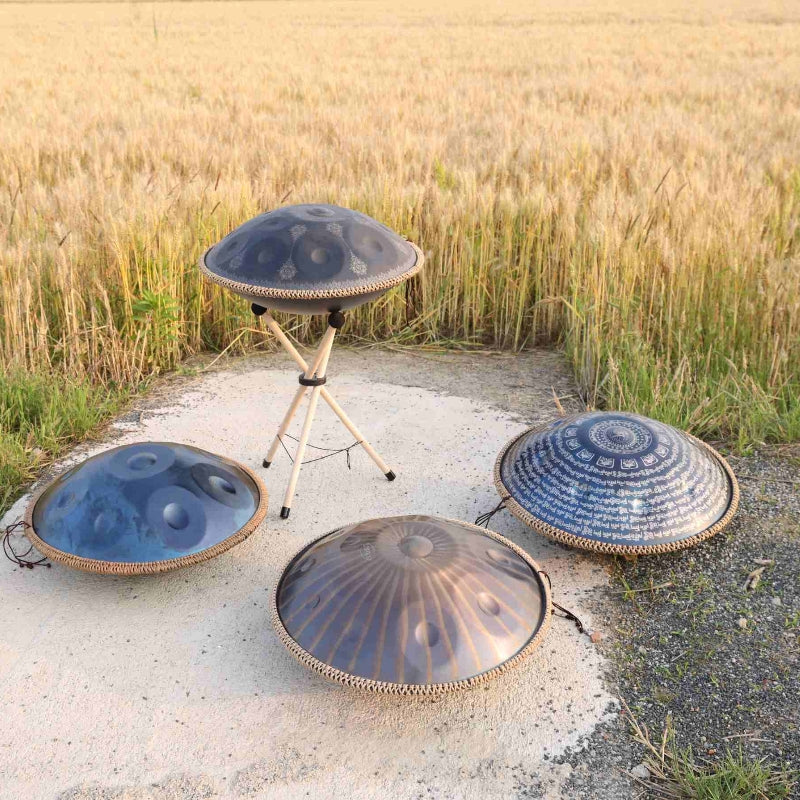 Four handcrafted handpans with engraved designs displayed outdoors near a wheat field, showcasing unique finishes and scales for meditation and music therapy.