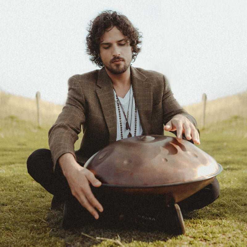 Male musician playing handpan on a grassy meadow outdoors.