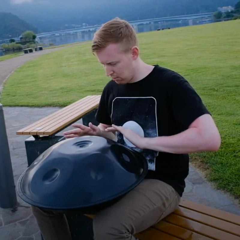 Male artist playing handpan drum outdoors while seated on a bench, creating soothing melodic rhythms in a natural setting.