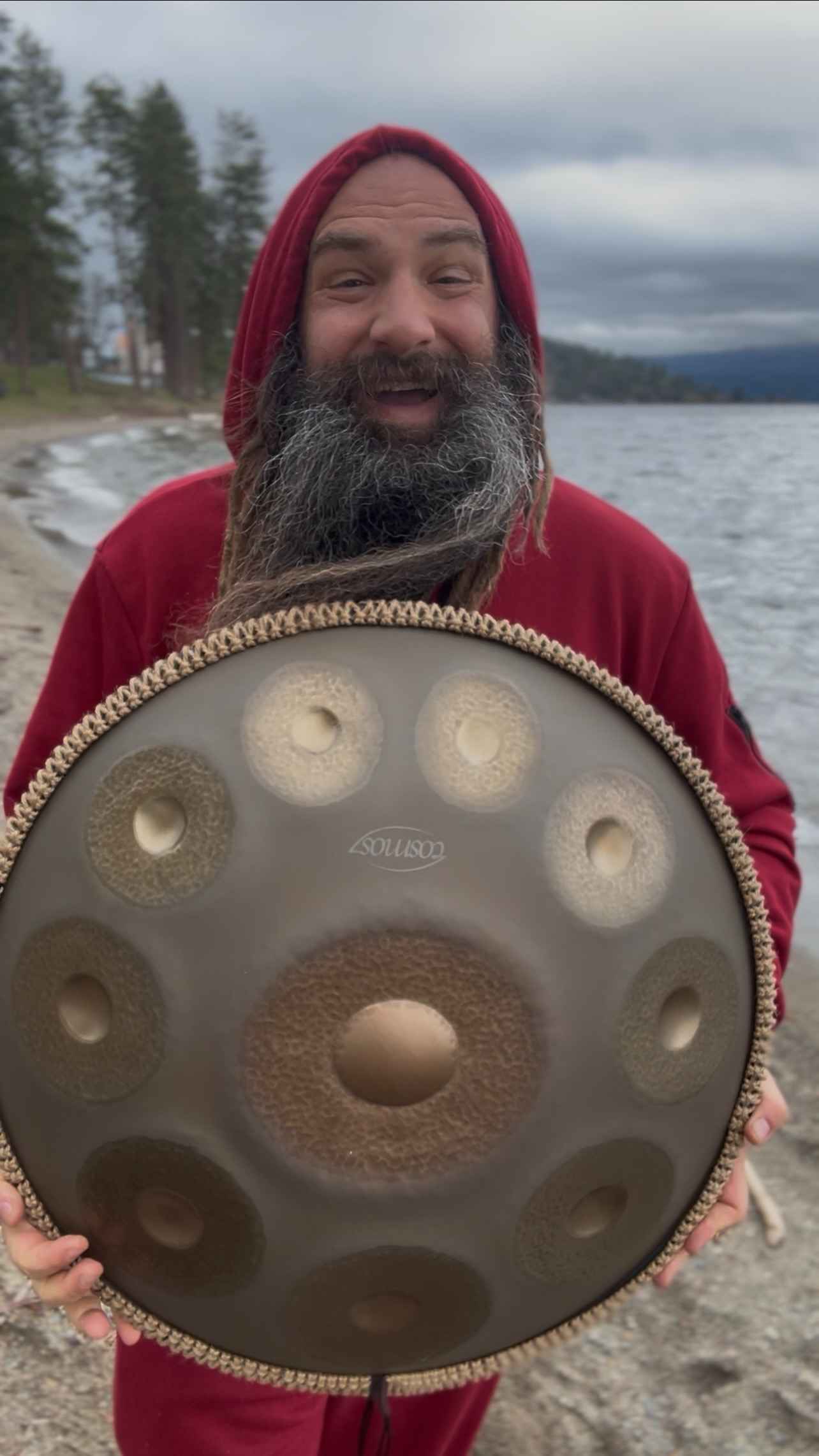 Person holding a large handpan on a beach with water and trees in the background