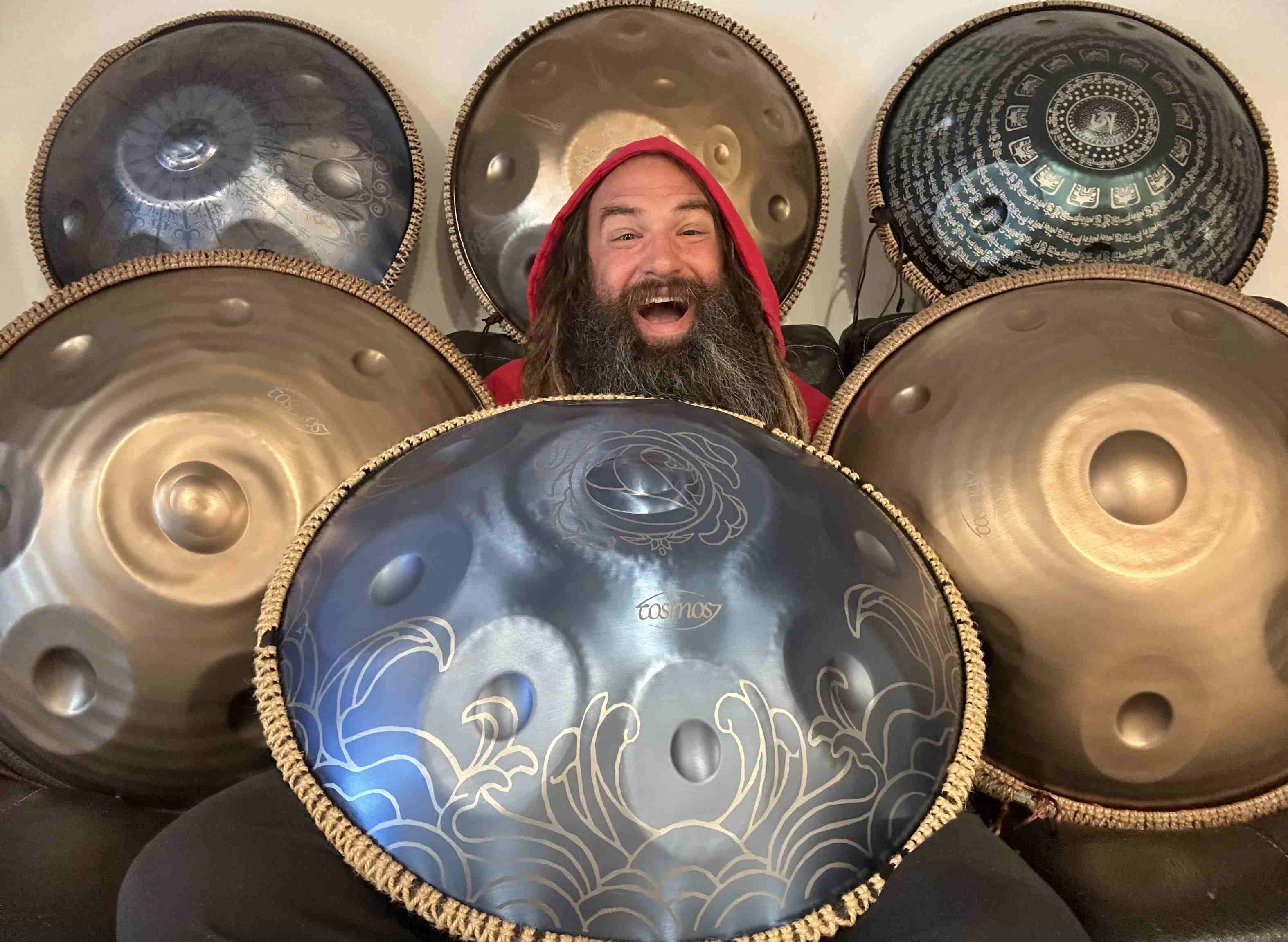 Smiling musician holding a blue engraved Cosmos handpan drum instrument surrounded by multiple handpans in different designs.